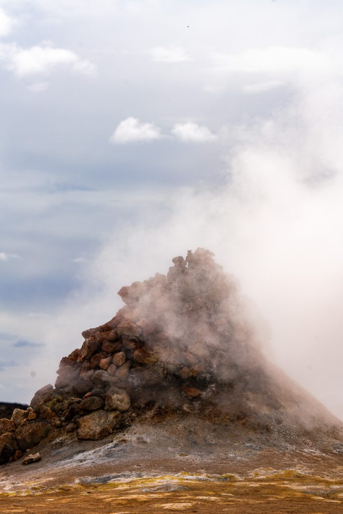 Een heuvel van dampende rotsen rijst op uit het dorre landschap van IJsland, met witte stoomwolken die naar een bewolkte hemel drijven. De grond lijkt droog en gelig rond de formatie, wat erop wijst dat er weinig ruimte is voor groei in dit ruige terrein.