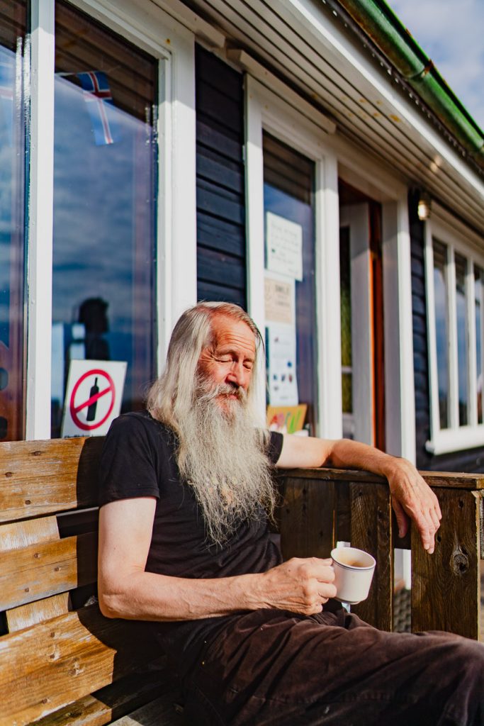 Een oudere man met lang wit haar en een baard zit op een houten bankje buiten een gebouw in IJsland, met een witte mok in zijn hand. Het zonlicht baadt hem terwijl hij geniet van het vredige moment. Op het raam erachter is een bordje "niet roken" te zien.