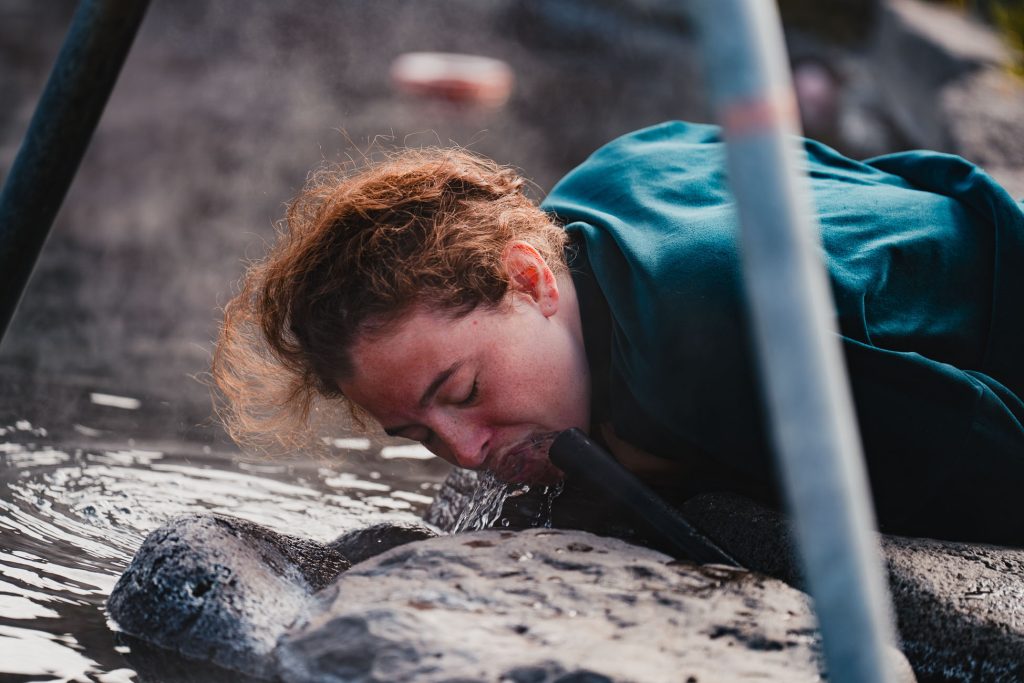 Een persoon met krullend bruin haar buigt voorover om water te drinken uit een pijp omringd door rotsen buiten in IJsland. De scène ziet er rustiek en natuurlijk uit, met zachte verlichting die het gezicht van de persoon benadrukt en een gevoel van pure groei in de natuur.