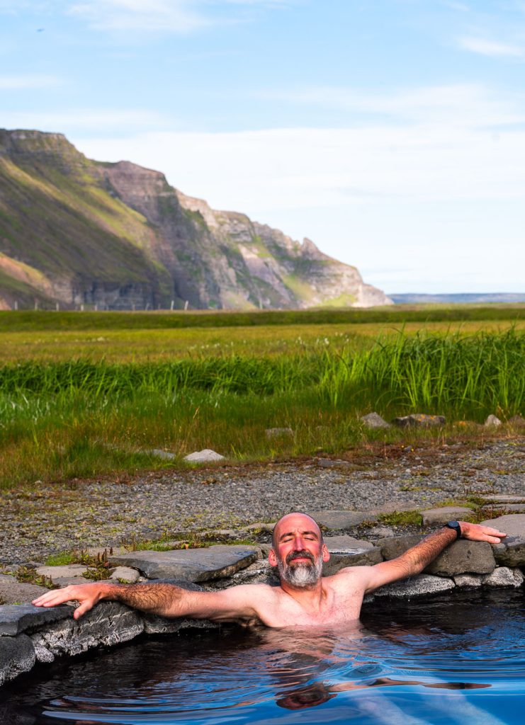 Een man ontspant met uitgestrekte armen in een natuurlijke warmwaterbron in IJsland, omgeven door gras en rotsachtig terrein, met groene bergen en een blauwe lucht die de wilde schoonheid waar het avontuur groeide verraadt.