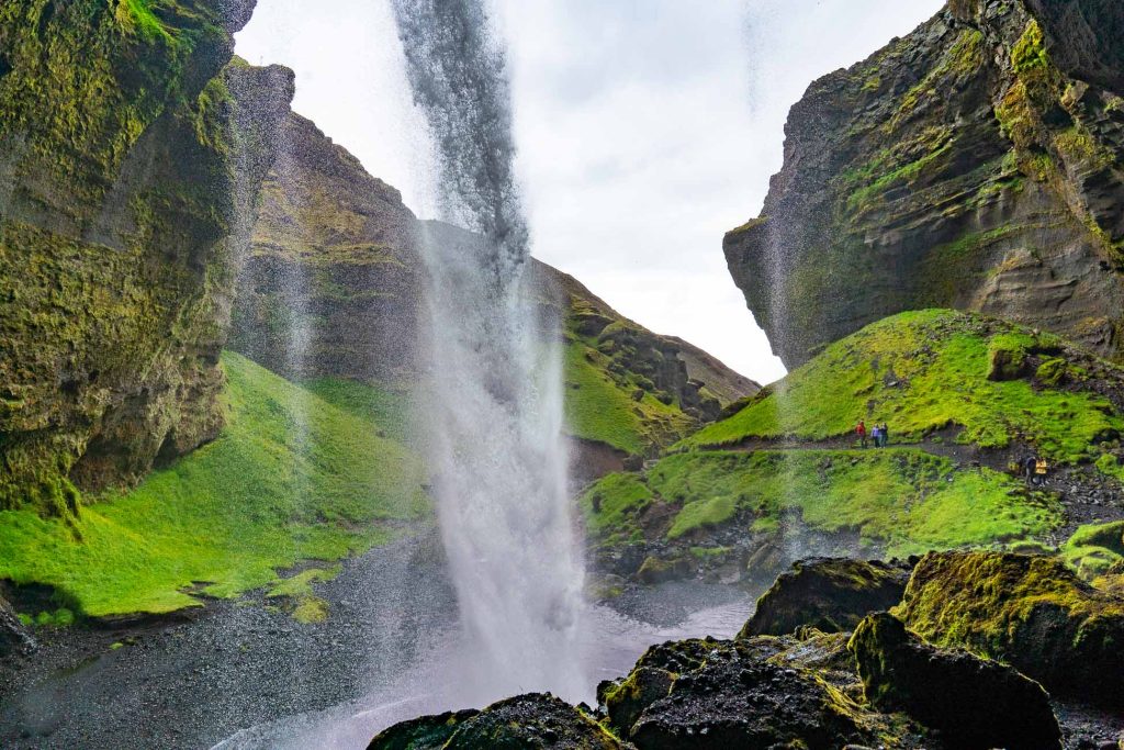 Een uitzicht vanachter een hoge waterval tussen met mos bedekte kliffen, met wandelaars langs een weelderig pad - een iconisch gezicht dat je kunt tegenkomen tijdens een 7-daagse roadtrip met Iceland Nomads door de adembenemende landschappen van Roadtrip IJsland.