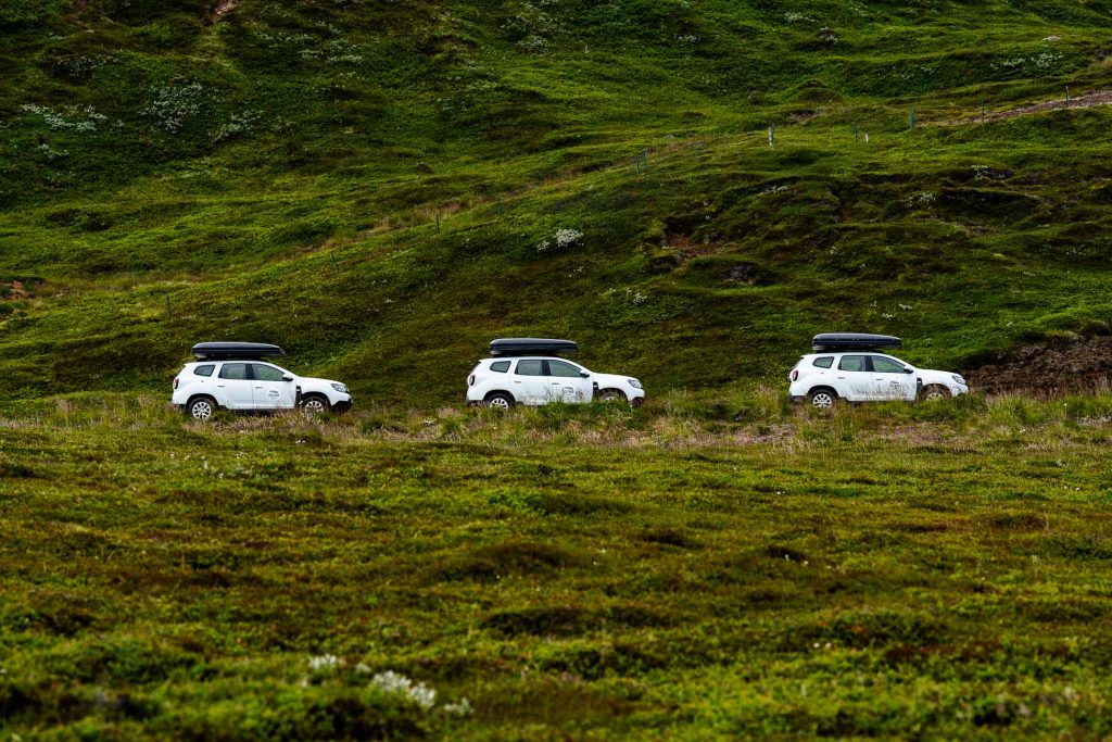 Drie witte SUV's met dakkoffers rijden in een lijn door een weelderig, groen, graslandschap met glooiende heuvels-een perfecte scène van een 7-daagse roadtrip met Iceland Nomads door het adembenemende Roadtrip IJsland.