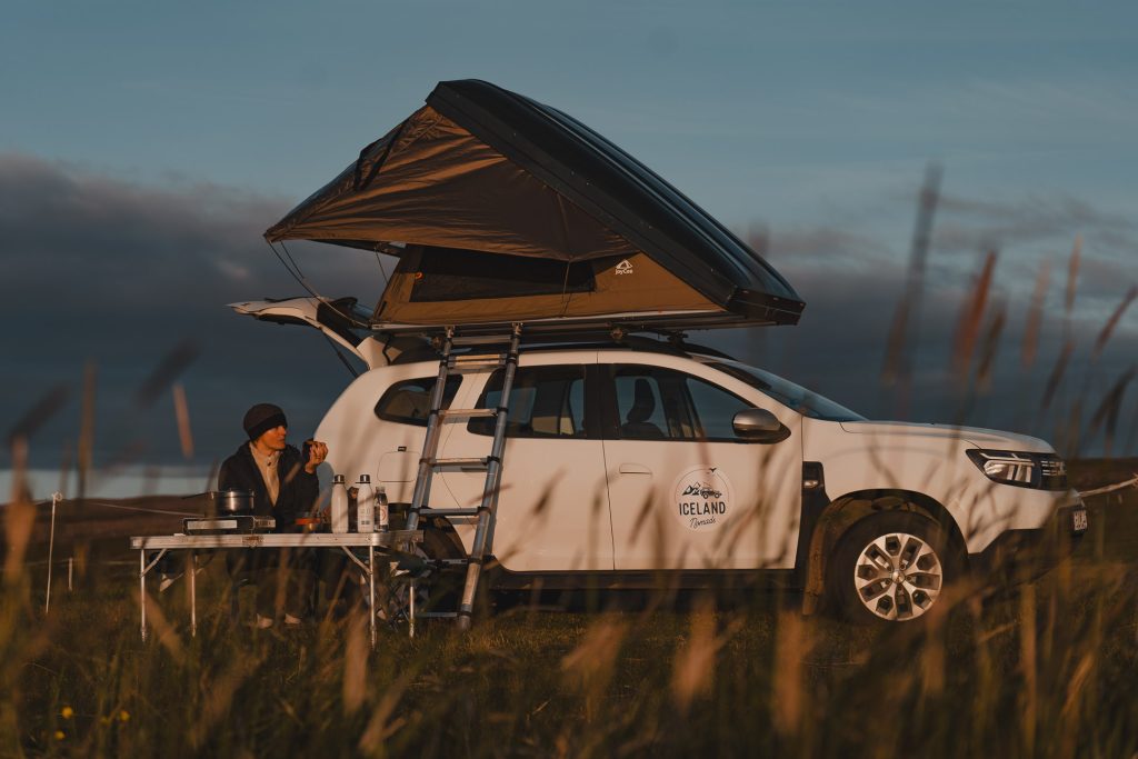 Een persoon zit aan een campingtafel naast een witte SUV met daktent en geniet van de zonsondergang in een grasveld. Kampeerspullen en het voertuig met het opschrift "Iceland Nomads" duiden op een epische 7-daagse roadtrip door de schilderachtige landschappen van Roadtrip IJsland.