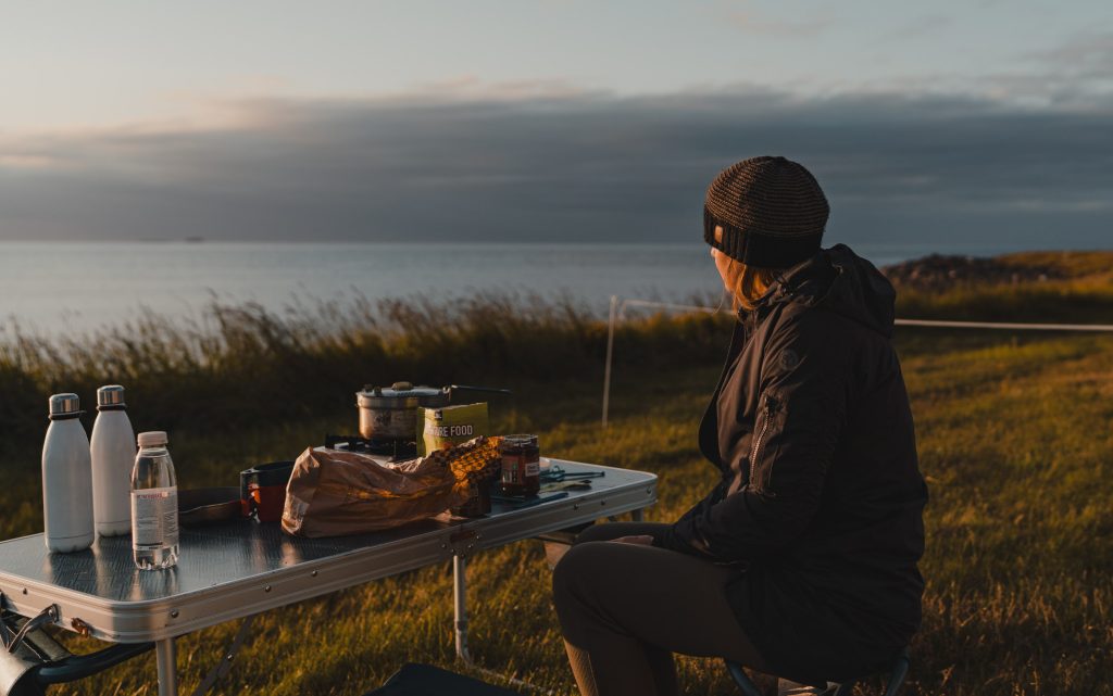 Een persoon in een donkere jas en muts zit aan een campingtafel met eten en waterflessen, genietend van het uitzicht bij zonsondergang over kalm water en grasvelden - een ideale scène van een 7-daagse roadtrip met Iceland Nomads.