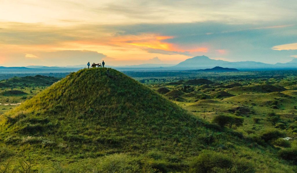Een kleine groep staat op de top van een met gras begroeide heuvel bij zonsondergang in Afrika, met een uitgestrekt landschap en bergen in de verte op de achtergrond onder een kleurrijke hemel.