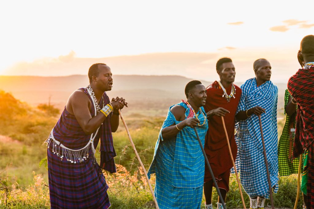 Vijf Maasai mannen in traditionele kleurrijke gewaden en kralenwerk staan samen buiten bij zonsondergang in Tanzania, met houten staven in de hand, omringd door grasvlakten en heuvels in Afrika in de verte.
