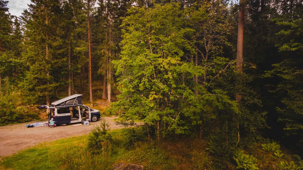 Een camper met het dak omhoog staat geparkeerd op een onverharde weg in een dicht bos in Zweden, met buiten kampeerspullen klaargezet. Weelderige groene bomen omringen de vredige, openluchtscène - perfect om het naturisme te midden van de natuur te omarmen.