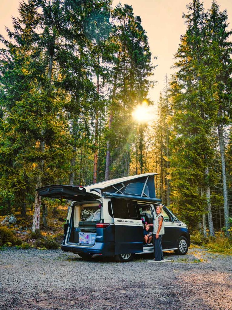 Een camper met open deuren staat geparkeerd tussen hoge dennenbomen in Zweden. Twee mensen staan naast de bus en genieten van het naturisme en het zonlicht dat door het bos schijnt. Achterin de bus zijn kampeerspullen zichtbaar.