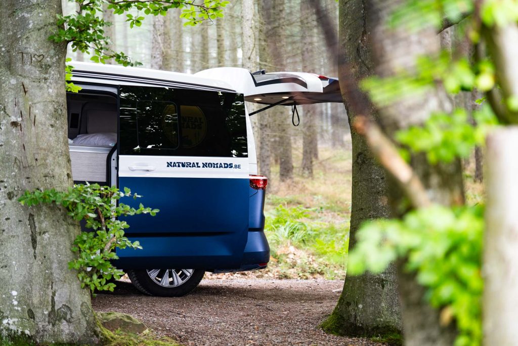 Een blauw-witte camper met "Natural Nomads" op de zijkant staat geparkeerd in een Zweeds bos. De achterklep van het busje staat open, het interieur is gedeeltelijk zichtbaar tussen de bomen en biedt een serene omgeving voor liefhebbers van naturisme.