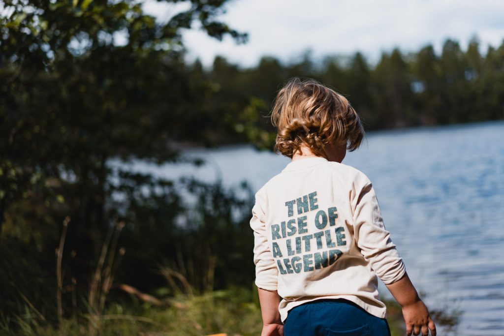 Een jong kind met blond haar staat bij een meer in Zweden en kijkt de andere kant op. Het kind draagt een shirt met de tekst "THE RISE OF A LITTLE LEGEND. Bomen en wazig water, perfect voor naaktrecreatie, staan op de achtergrond.