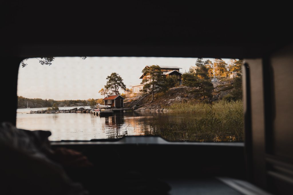 Zicht vanuit een voertuig op een vredig meer in Zweden met houten hutten omringd door bomen, weerspiegeld in het kalme water bij zonsondergang.