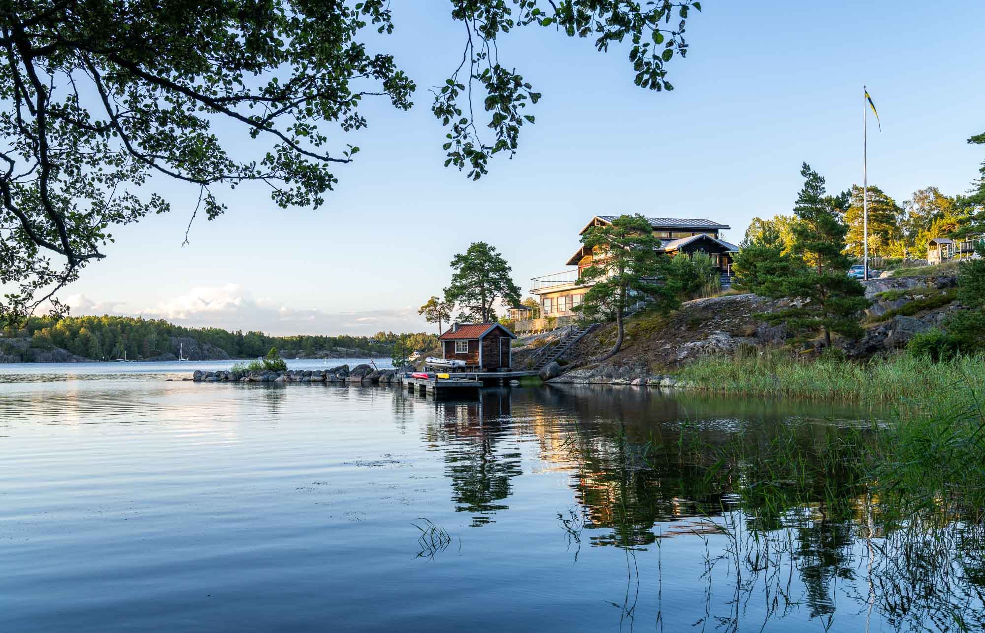 Een vredig tafereel aan een meer in Zweden, met huizen genesteld tussen bomen aan een rotsachtige oever, een steiger die uitsteekt in kalm water en een Zweedse vlag die wappert op de achtergrond onder een heldere hemel.