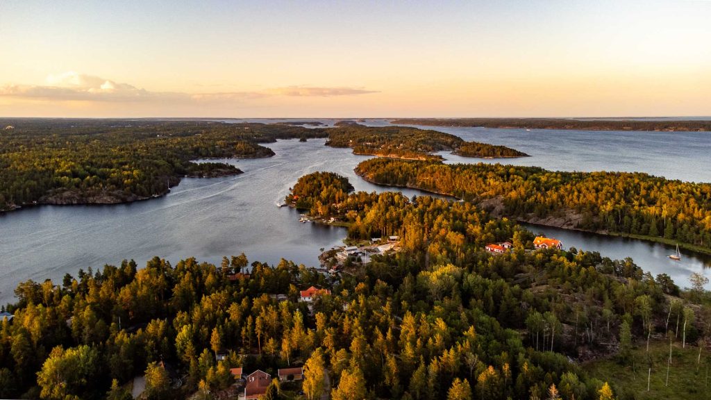 Luchtfoto van een serene archipel in Zweden met dichte bossen en verspreide huizen langs de waterkant bij zonsondergang, omringd door rustig blauw water en eilanden in de verte-een perfecte omgeving voor vredige naaktreizen.