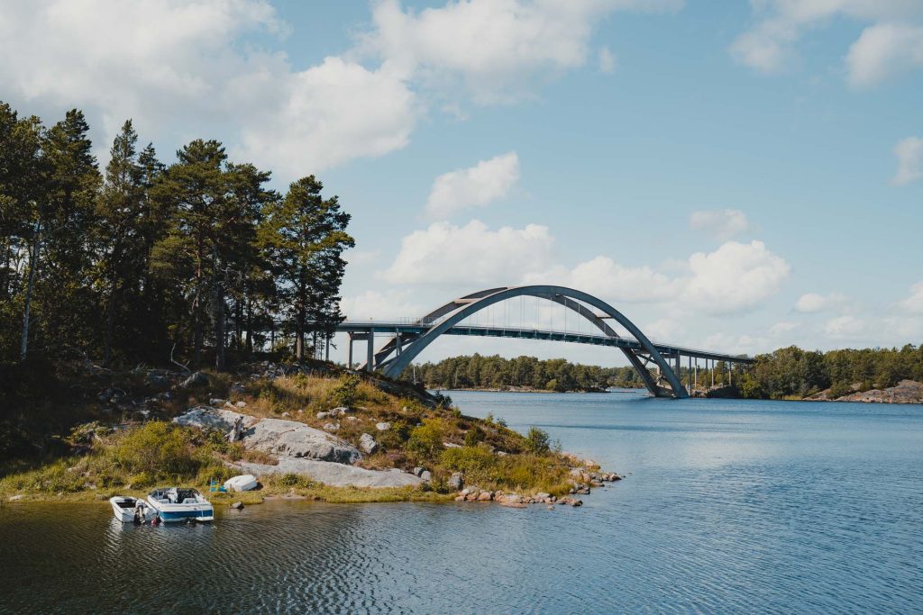 Een schilderachtig uitzicht op een moderne boogbrug in Zweden over een brede rivier, omringd door bomen en rotsachtige oevers, met een kleine boot aangemeerd aan de waterkant onder een gedeeltelijk bewolkte hemel.