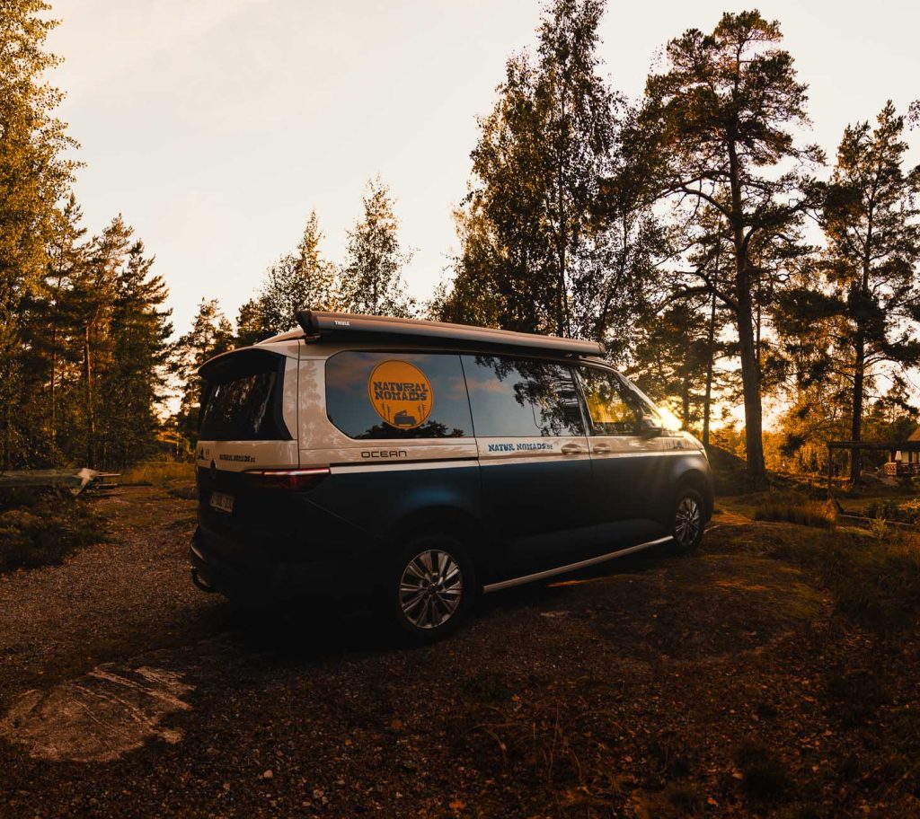 Een camper met een "Nationaal Park" logo staat geparkeerd op een grindvlakte omringd door bomen bij zonsondergang in Zweden, met warm zonlicht dat door het gebladerte filtert, perfect voor wie houdt van naturisme en één zijn met de natuur.