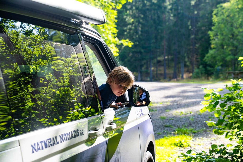 Een kind leunt uit het raam van een geparkeerd busje in een bosrijk gebied in Zweden. Zonlicht filtert door de bomen en op de zijkant staat "NATUURLIJK NOMADS BE" geschreven. Het kind kijkt nieuwsgierig naar buiten, de geest van naaktrecreatie omarmend.