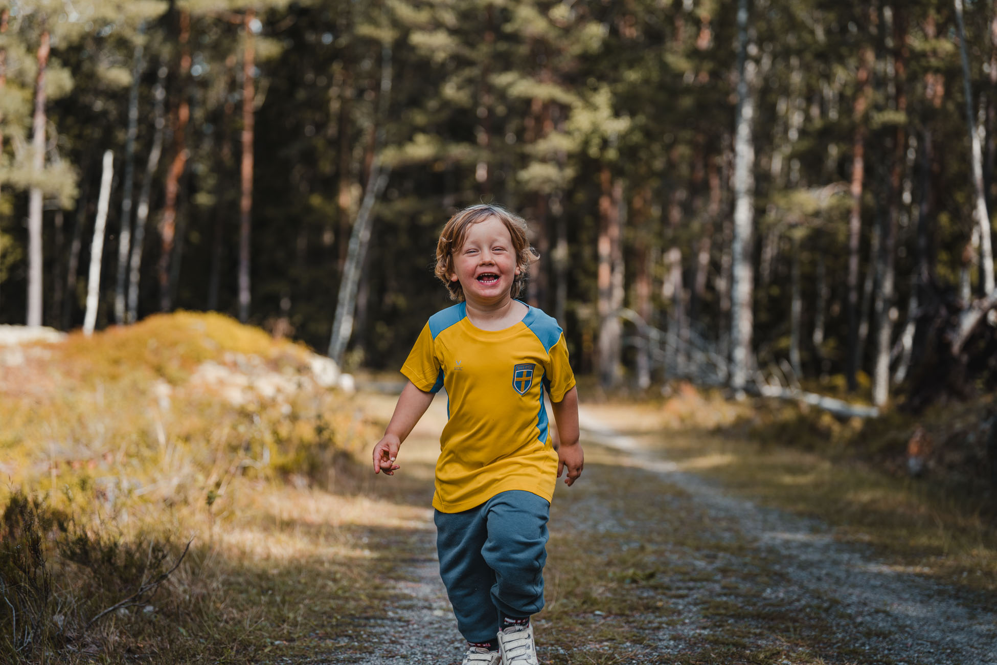 Een jong kind in een geel shirt met een blauw embleem rent vrolijk over een bospad in Zweden, omringd door bomen en groen, op een zonnige dag.