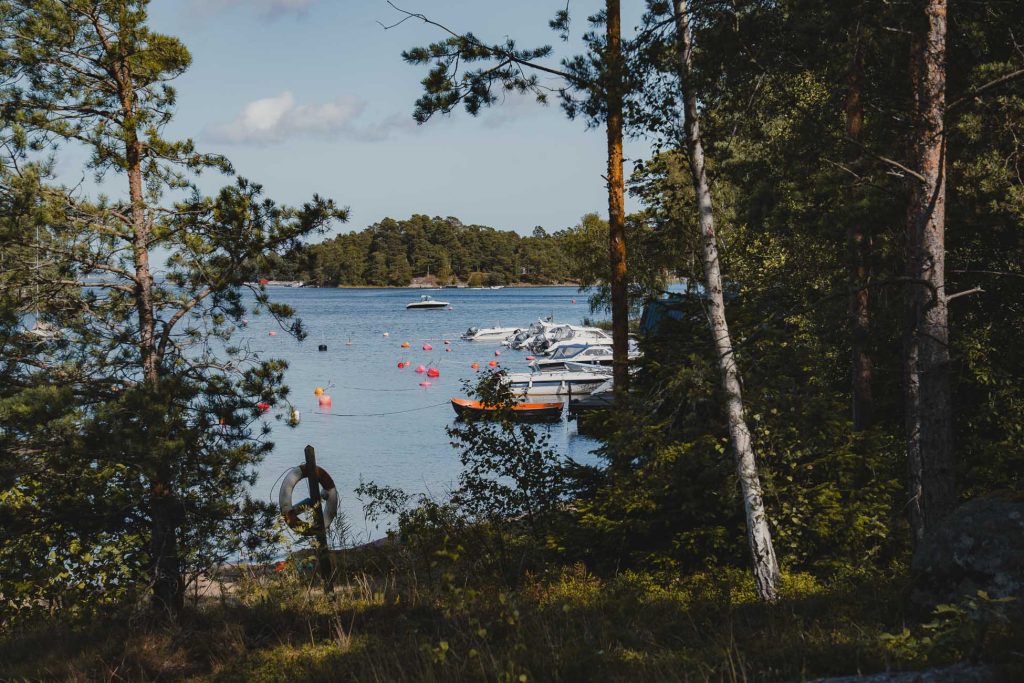 Zicht op een rustig meer in Zweden omlijst door bomen, met verschillende boten aangemeerd aan een pier, rode boeien drijvend in het water en een bebost eiland zichtbaar op de achtergrond onder een blauwe lucht met verspreide wolken.