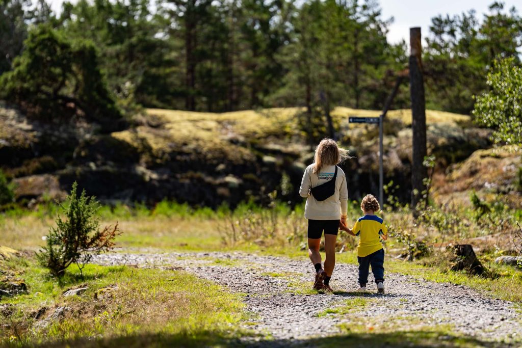 Een volwassene en een kind lopen hand in hand over een grindpad in een zonnig, groen bosgebied in Zweden. De volwassene draagt een rugzak en beiden zijn van achteren te zien terwijl ze het naturisme-vriendelijke pad met bomen en gras verkennen.