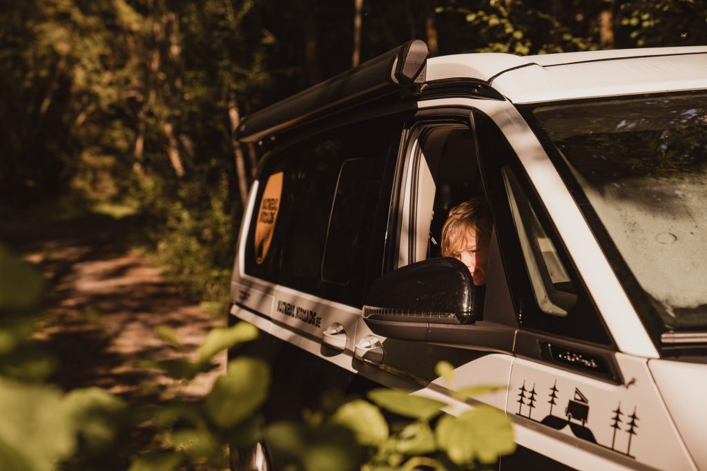 Een kind kijkt naar buiten vanuit het open raam van een geparkeerde camper op een zonovergoten bosweg in Zweden, met groen en bomen die het tafereel omringen.