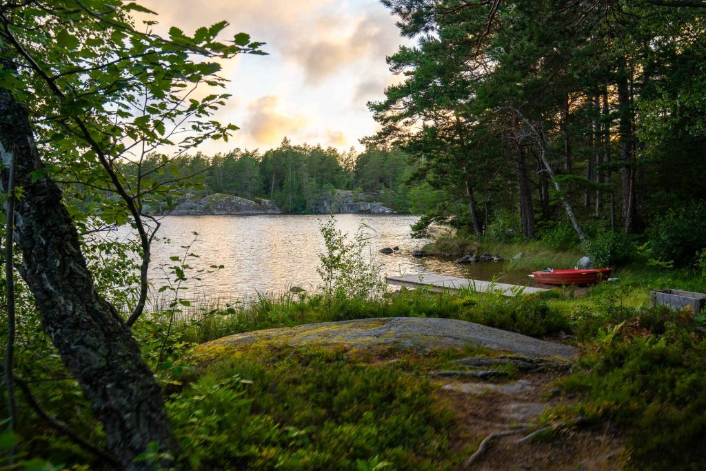 Een vredig tafereel aan een meer in Zweden met bomen en rotsen op de voorgrond, een kleine aanlegsteiger en rode roeiboot rechts, en een rustig meer omringd door bossen onder een gedeeltelijk bewolkte hemel bij zonsondergang.