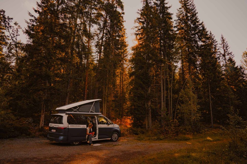 Een persoon zit buiten een camper met een verhoogd dak, geparkeerd tussen hoge dennenbomen in een Zweeds bos, genietend van de warmte van het zonlicht dat door de takken filtert - perfect om het naturisme in het mooie Zweden te omarmen.