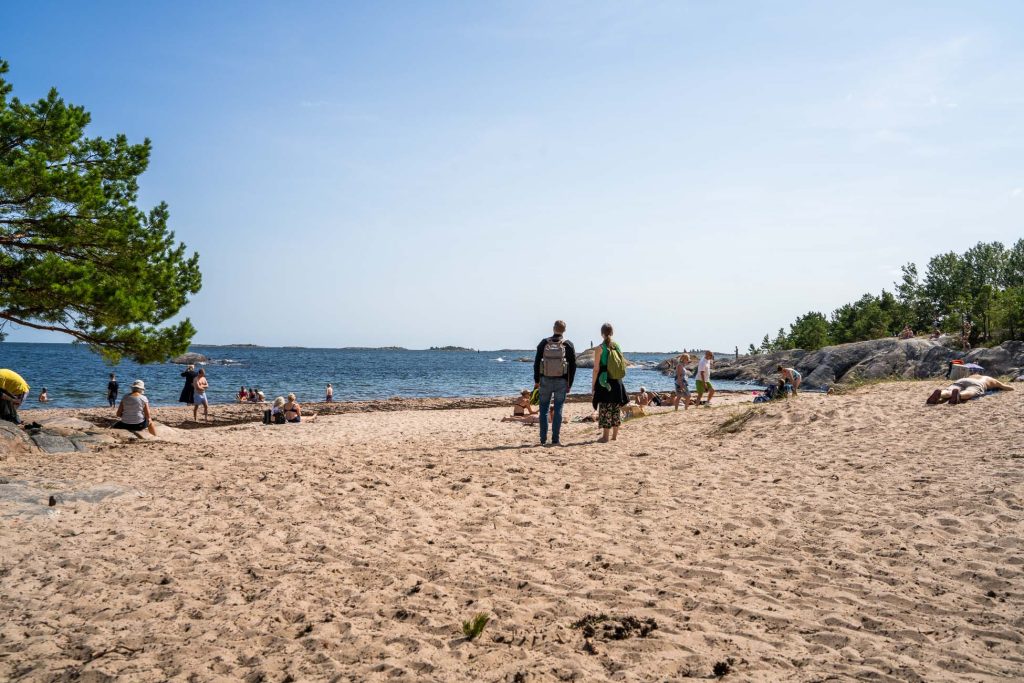 Mensen relaxen op een zandstrand aan zee in Zweden. Sommigen genieten van naturisme, liggend op handdoeken terwijl twee mensen met rugzakken naar het water lopen. Bomen en rotsen omringen het strand onder een helderblauwe hemel.