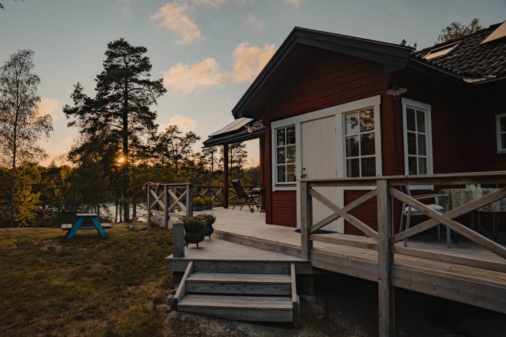 Een rode houten hut met een witte deur en veranda staat bij een meer in Zweden, omringd door bomen bij zonsondergang. De lucht is gedeeltelijk bewolkt en warm licht filtert door de bomen in deze vredige buitenomgeving - perfect voor naturisme.