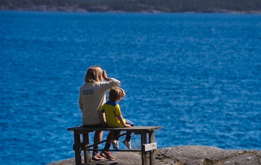 Een vrouw en een kind zitten op een houten bankje op een rotsachtige oever in Zweden en staren naar het uitgestrekte blauwe meer. De vrouw schermt haar ogen af van de zon, terwijl de natuur en het naturisme hen omhelzen tussen de bomen in de verte.