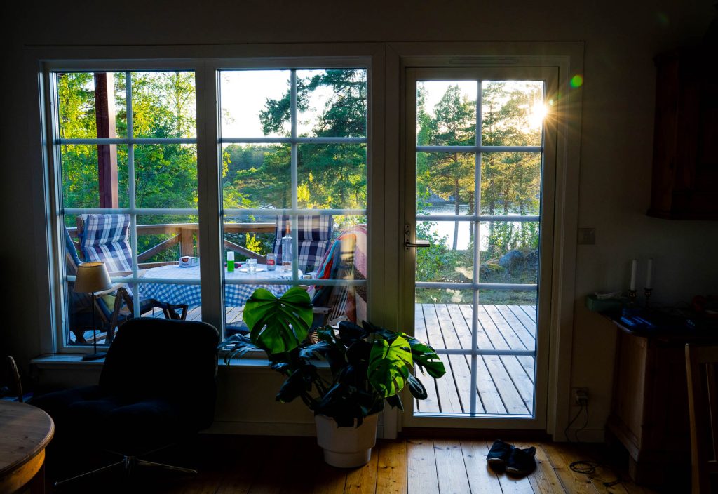 Een zonovergoten patio met uitzicht door glazen deuren, met buitenstoelen rond een blauwgeruite tafel, straalt een Zweeds geïnspireerde charme uit. De gezellige kamer binnen is omgeven door groen en heeft kamerplanten, een stoel en schoenen.