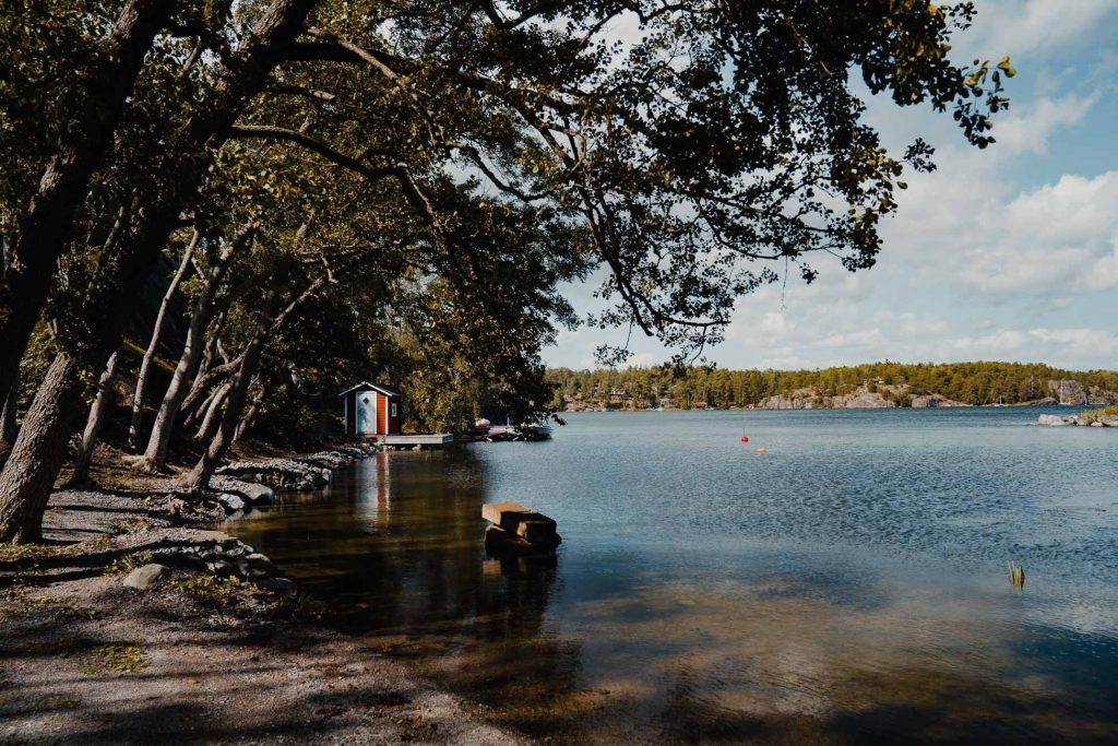 Een kalme meeroever in Zweden met bomen die over het water leunen, een klein rood-wit boothuis helemaal links en rode boeien die in het water drijven onder een halfbewolkte hemel - perfect voor een sereen blootje-moment.