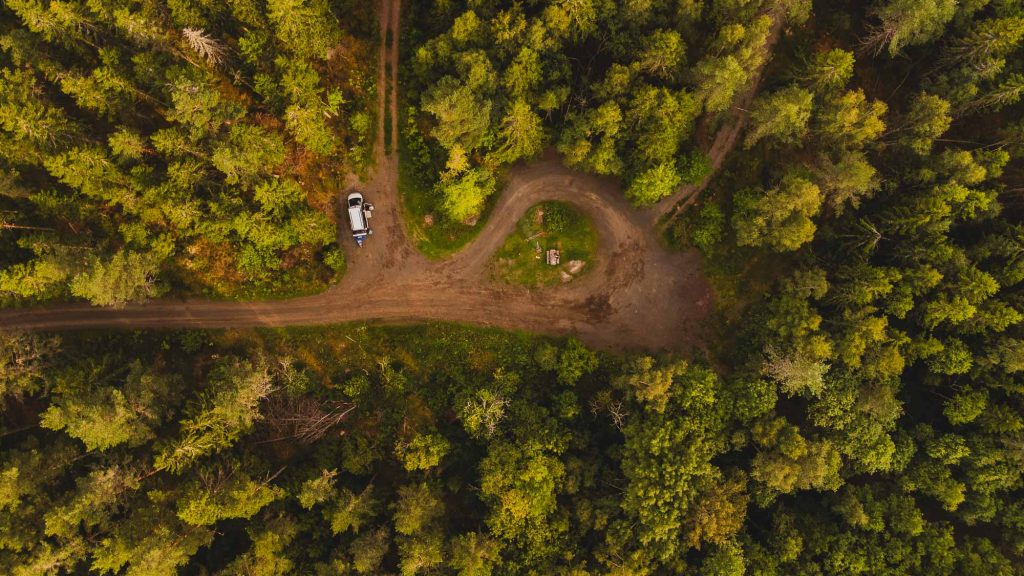 Luchtfoto van een open plek in het bos met een onverharde weg die kronkelt tussen dichte groene bomen in Zweden; een wit voertuig staat geparkeerd bij de weg, perfect voor wie wil genieten van bloot recreëren in warm, gouden zonlicht.