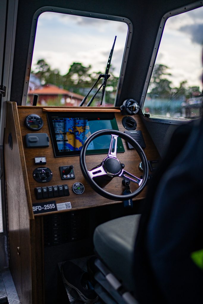 Een close-up van een houten bedieningspaneel van een boot met een stuurwiel, een elektronisch navigatiescherm met een kaart van Zweden, verschillende knoppen en een voorruit met een wazige buitenwereld.