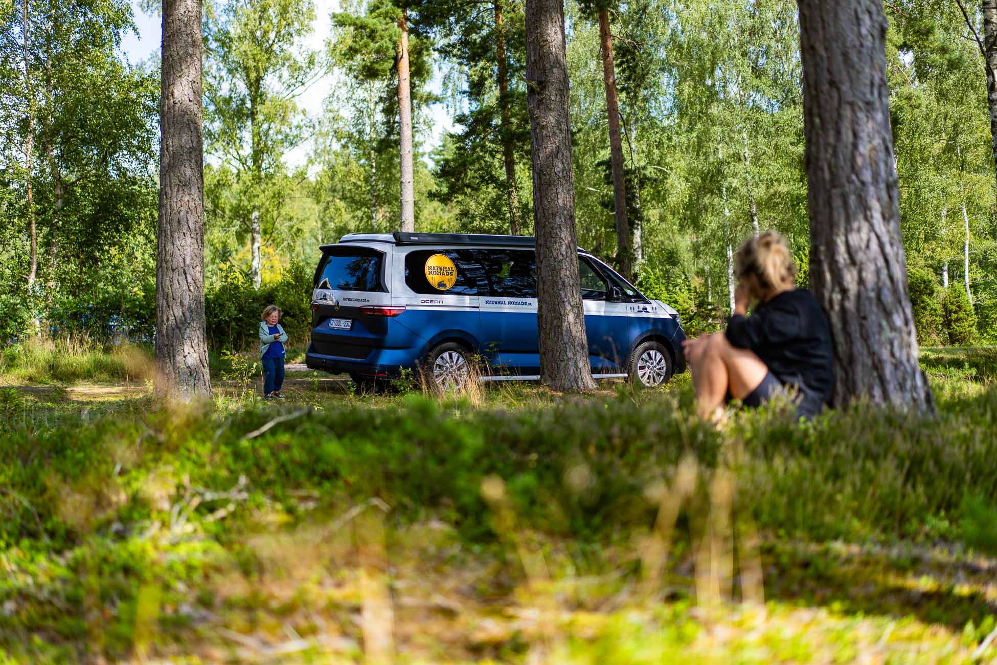 Een blauwe camper staat geparkeerd tussen hoge bomen in een Zweeds bos. Een kind staat bij het busje terwijl een volwassene op het gras op de voorgrond zit en naar het kind kijkt. Zonlicht filtert door de bomen en creëert een serene naturistische sfeer.