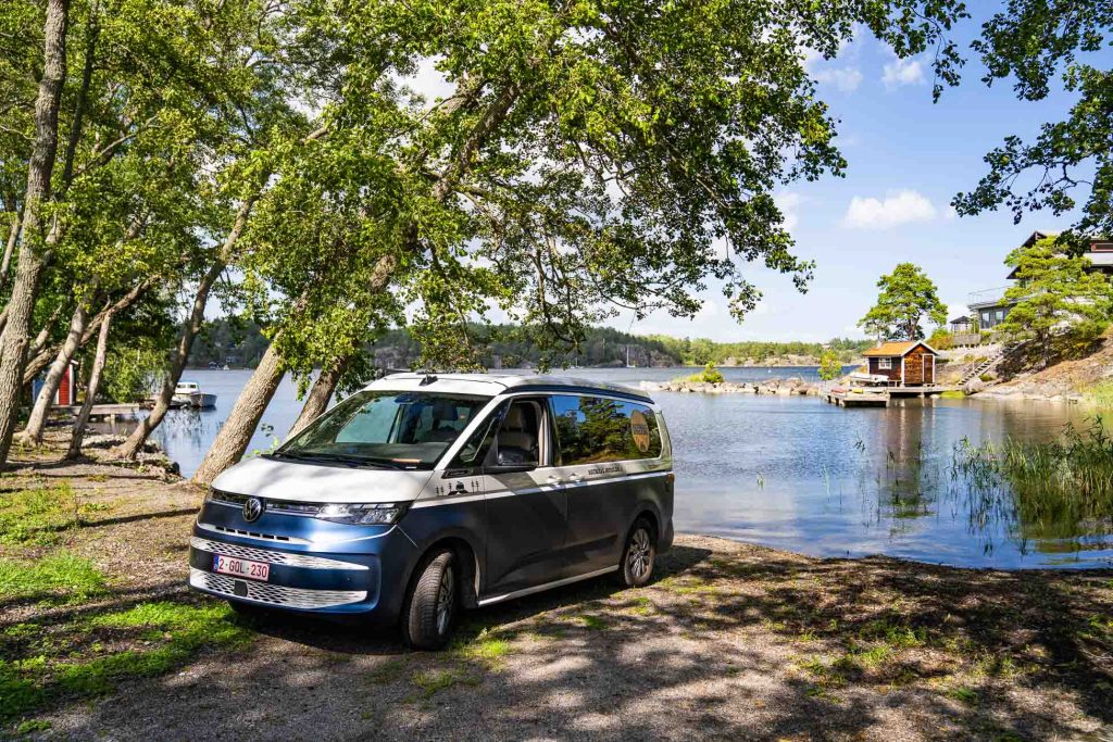 Een blauw-wit busje staat geparkeerd aan een rustig meer in Zweden, omringd door bomen en hutten. De helderblauwe lucht draagt bij aan het vredige tafereel, met water, groen en houten huizen op de achtergrond.