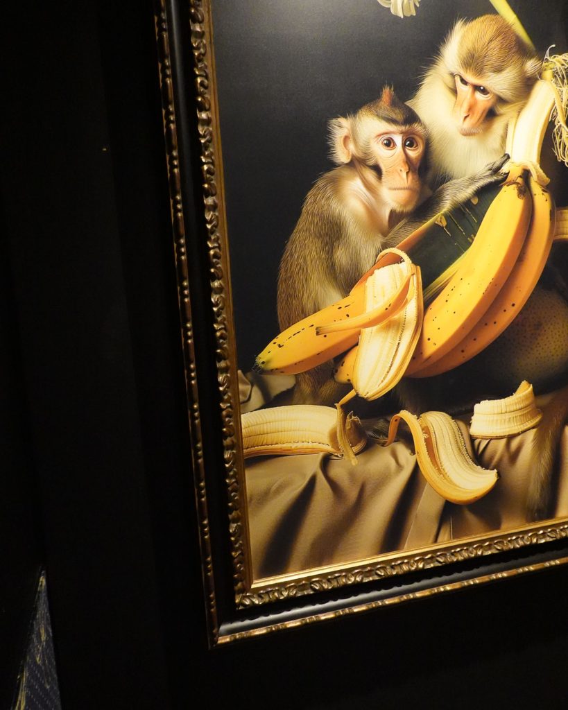 Een ingelijst schilderij toont twee apen die samen zitten, omringd door gepelde bananen op een gedrapeerde doek, tegen een donkere achtergrond. De sierlijke gouden lijst geeft het kunstwerk een klassiek tintje dat doet denken aan galeries in Hasselt.