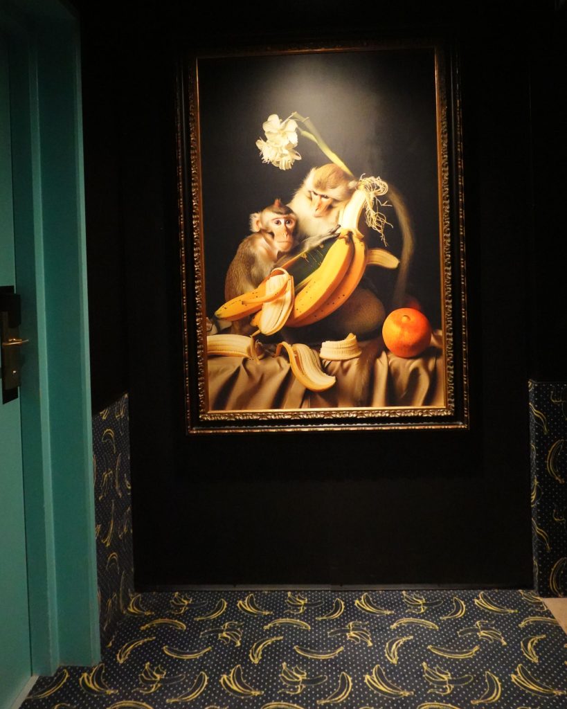 Een ingelijst schilderij van twee apen met bananen, een sinaasappel en een bloem hangt aan een zwarte muur in Hasselt. Op de vloer ligt een blauw tapijt met gele bananenpatronen en links is een deel van een turkooizen deur zichtbaar.