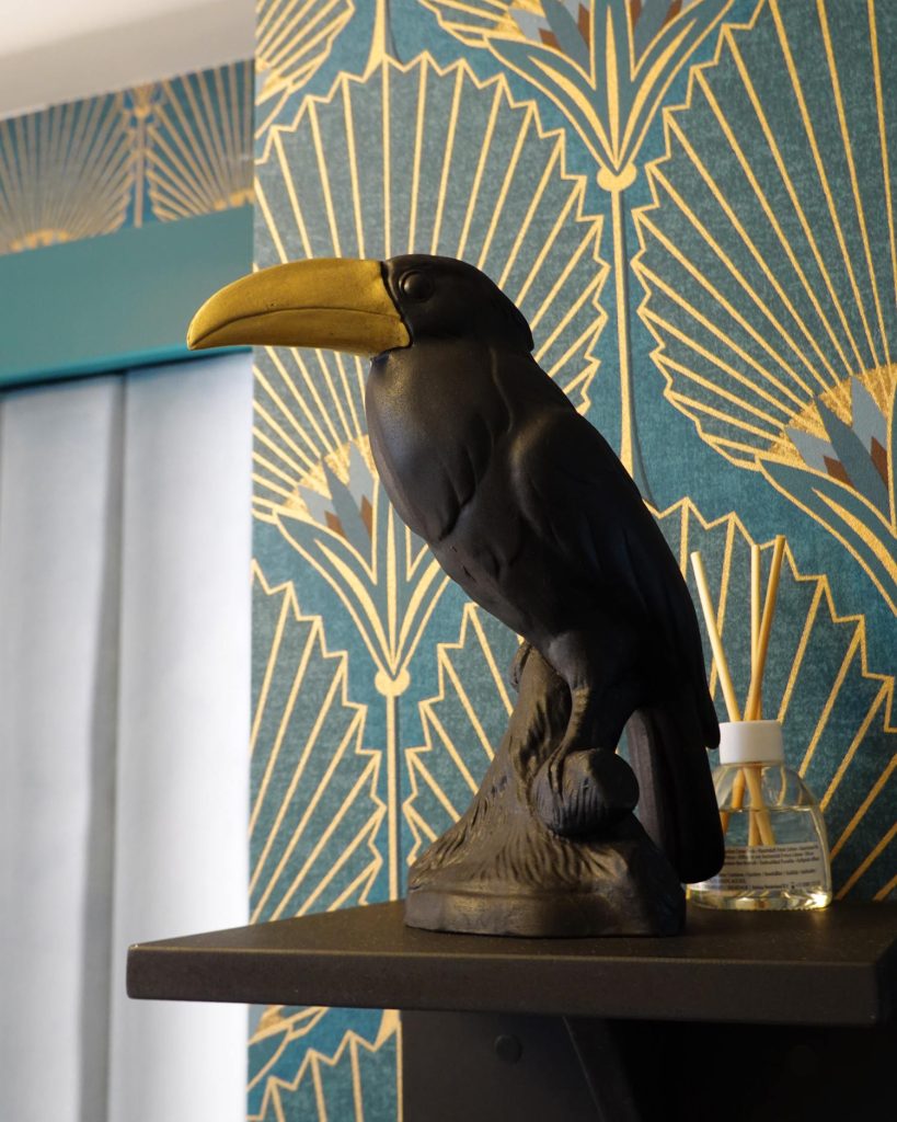 Een zwart met gouden toekan beeldje staat op een plank voor blauw met goud gedessineerd behang, wat een vleugje Hasseltse charme toevoegt. Een kleine fles met rietverspreiders staat op de achtergrond.