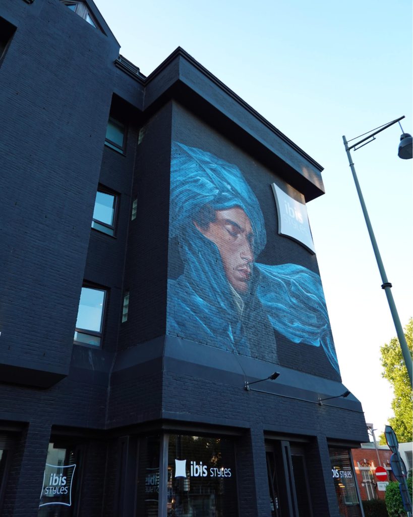 Een muurschildering op de buitenmuur van een donker gebouw in Hasselt toont een persoon met gesloten ogen, gedrapeerd in een vloeiende blauwe stof. Het gebouw herbergt een Ibis Styles hotel, zoals de bewegwijzering aangeeft, en wordt verlicht door daglicht.