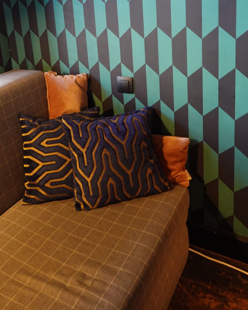Een grijs geruite sofa met twee kussens met donkerblauw en goudpatroon en twee oranje kussens met structuur zit voor een groenblauwe en zwarte muur met geometrisch patroon, wat een vleugje moderne stijl brengt in elke Hasseltse woning.
