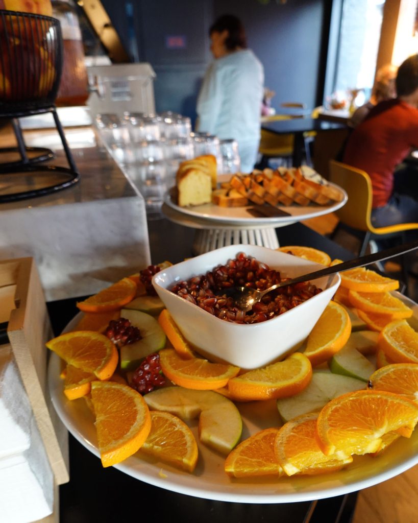 Een schaal met gesneden sinaasappels en appels omringt een schaal met granaatappelpitjes. Op de achtergrond genieten gasten in een café in Hasselt van gesneden cake op een standaard, met glazen in de buurt, wat een gezellige en uitnodigende sfeer creëert.