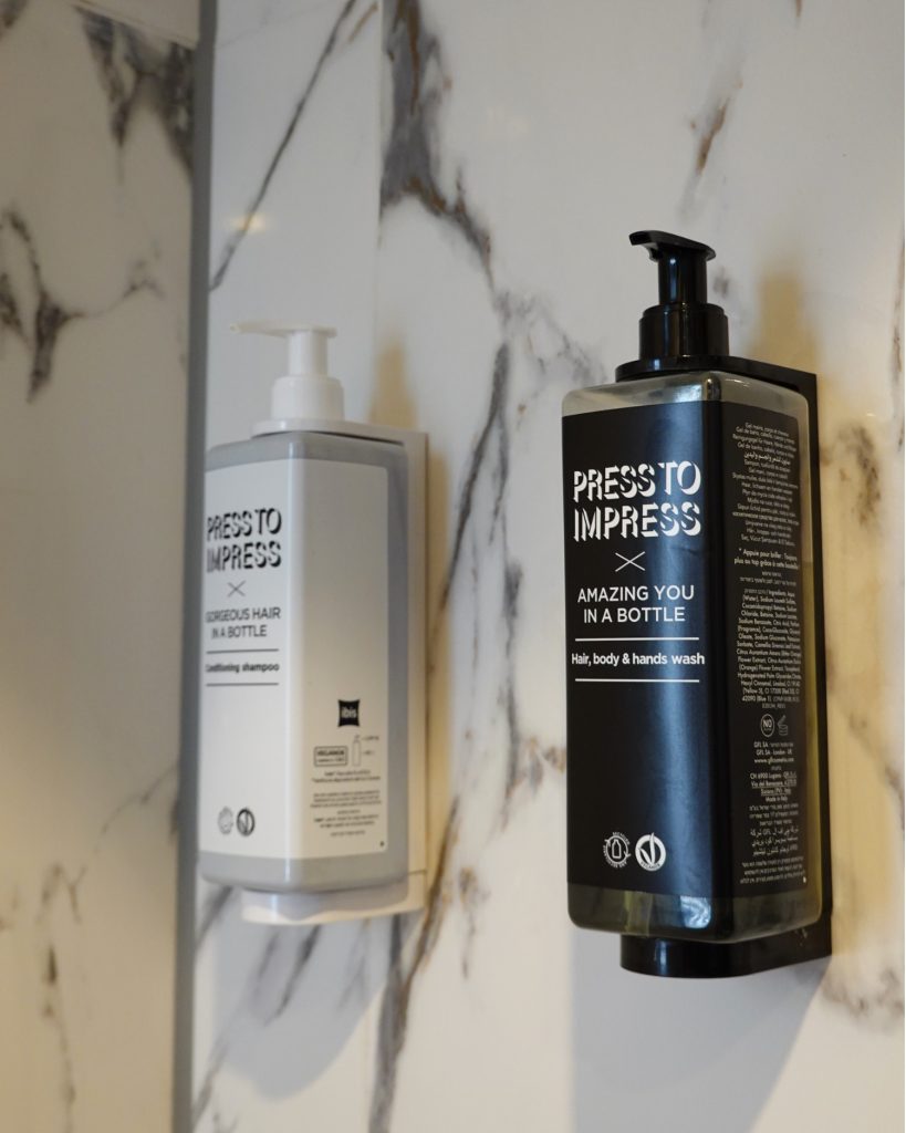 Twee wandflessen met het opschrift "Press To Impress", een witte en een zwarte, zijn bevestigd aan een met marmer betegelde badkamermuur in Hasselt. De witte fles is voor shampoo, de zwarte voor haar-, lichaams- en handwas.
