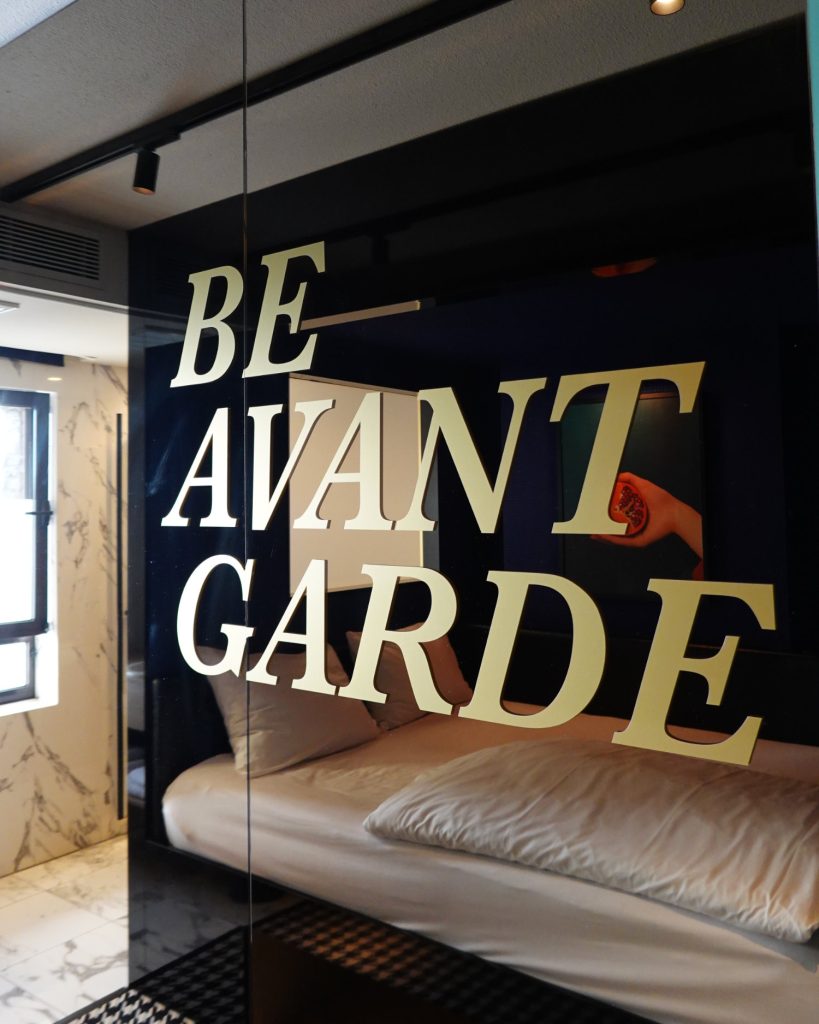 Grote beige letters met de tekst "BE AVANT GARDE" zijn gedrukt op een reflecterende glazen muur in een moderne, stijlvolle kamer met marmeren accenten en een geruite vloer - een vleugje Hasseltse flair ver van Antarctica.