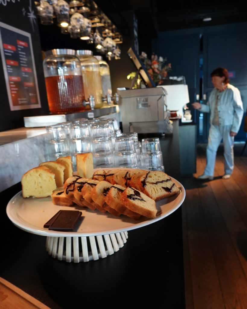 Een bord met plakjes cake en chocolade staat op een toonbank in een modern café in Hasselt. Glazen potten, bekers en menukaarten staan op de achtergrond, met een vrouw in lichte kleding die vlakbij loopt.