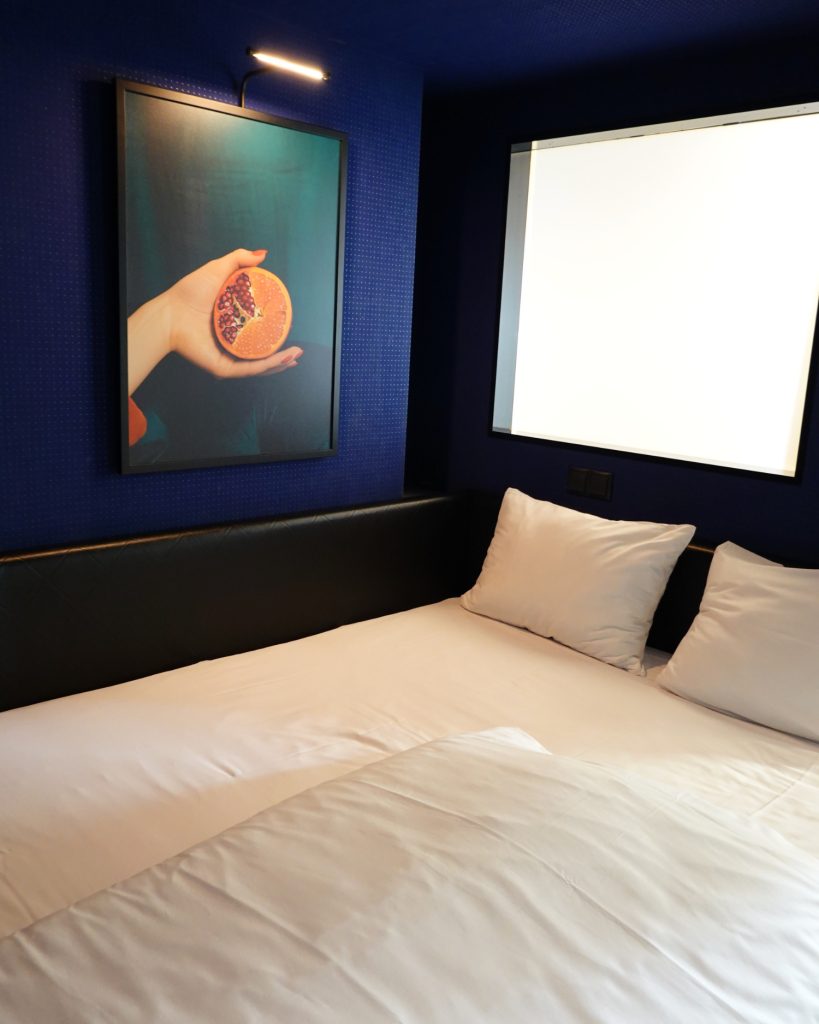 Een keurig opgemaakt bed met wit linnengoed staat naast een zwart hoofdeinde, een verlicht raam en een ingelijst kunstwerk met een hand die een halve grapefruit vasthoudt tegen een donkerblauwe muur - een gezellig tafereel dat doet denken aan een boetiekverblijf in Hasselt.
