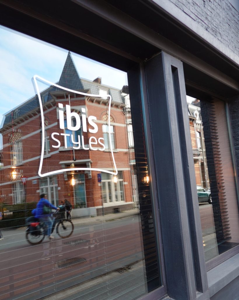 Een raam met het "ibis Styles" logo in Hasselt weerspiegelt een straatbeeld waarin een fietser onder een halfbewolkte hemel langs rode bakstenen gebouwen rijdt.