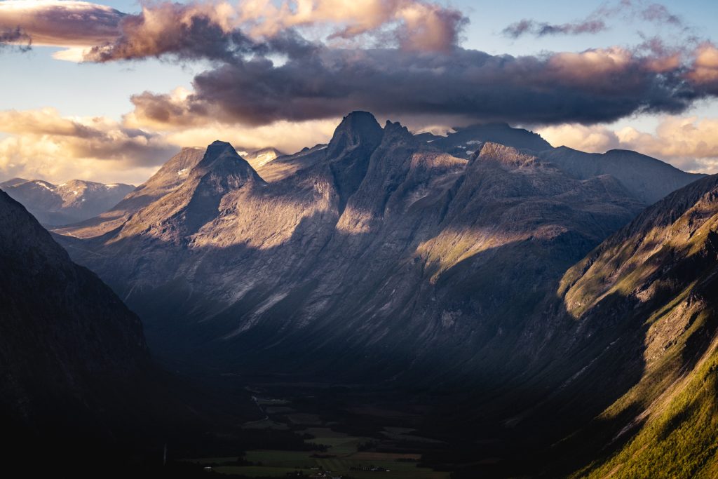 Dramatische bergtoppen en bergkammen in het fjordenland baden in gouden middernachtzon, terwijl donkere wolken schaduwen werpen over een diep dal eronder. Het ruige landschap wordt gekenmerkt door groene bossen en rotsachtige hellingen - echt Noorwegen Nomadengebied.