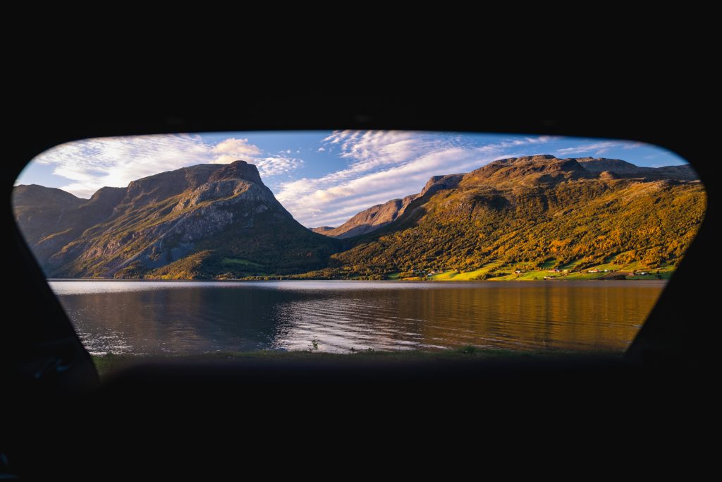 Uitzicht door een autoraam op een rustig fjordenmeer met weerspiegelingen, omringd door groene en gouden bergen onder een halfbewolkte middernachtzon-hemel - een echt Noorwegen Nomads tafereel.