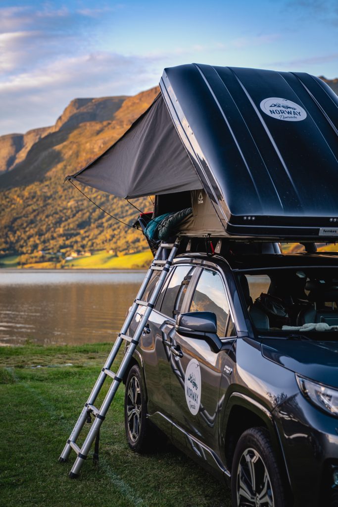 Een auto met daktent staat geparkeerd bij een rustige fjord in Noorwegen, met bergen op de achtergrond. Bij zonsondergang, onder de gloed van de middernachtzon, beklimt iemand de ladder en geniet van het rustige en schilderachtige uitzicht.
