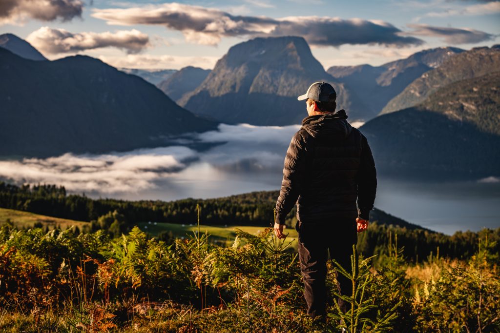 Een persoon met een jas en pet staat op een met gras begroeide heuvel, uitkijkend over een mistig fjord omringd door bergen onder een gedeeltelijk bewolkte hemel tijdens zonsopgang of zonsondergang-een sereen moment voor de Noorwegen Nomaden onder de middernachtzon.